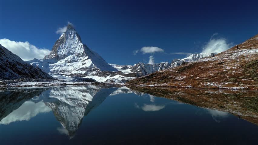 Riffelsee Lake mirrored reflection The Matterhorn famous mountain peak Zermatt Switzerland pan Swiss Alps aerial drone wind fog first fresh snow fall autumn winter blue sky morning Gornergrat railway