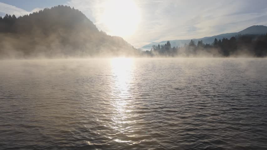 Morning mist rises over tranquil lake surrounded by mountains. Aerial shot of foggy lake at sunrise. Smoke on the water in morning sun