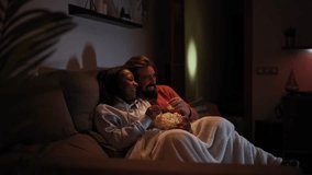 Side view of young multiracial couple relaxed watching late night movies lying on sofa at home eating popcorn. Two people together covered with a blanket in a dark living room enjoying a tv serie. - Powered by Shutterstock - Get 15% off with code: PIKWIZARD15