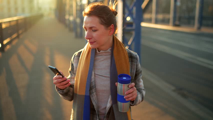 Woman walking in a frosty sunny morning, drinking coffee and using smartphone