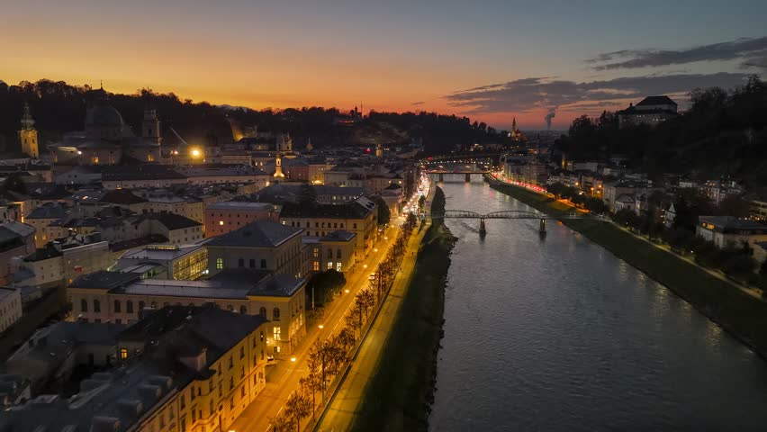Salzburg at sunset, Austria. Picturesque aerial view shows illuminated city buildings lining the Salzach River. Flying over Salzburg old town in the evening