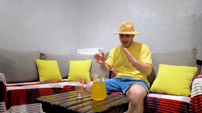 A young man drinks orange juice in an apartment. Drinking yellow lemonade juice from a transparent glass, slow motion.