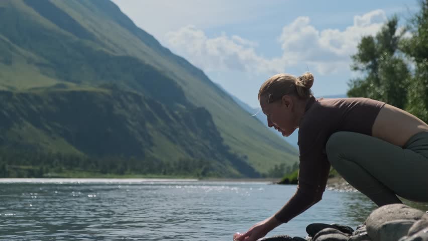 Happy adult woman in sport clothes washes face with clear mountain river water sitting on the stone on sunny day. Yoga classes during summer vacation in mountains.