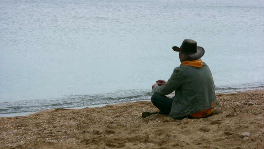 Wistful cowboy sits on a sandy beach and threw stones into the water