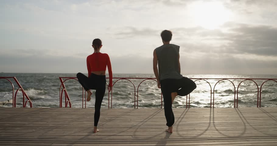 Happy couple guy and girl doing yoga and standing on one leg on the seashore in the morning in summer