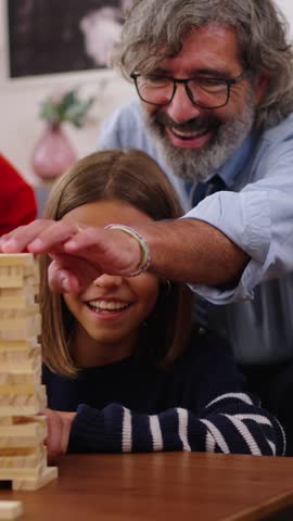 Vertical. Excited multi-generational family gathering playing Jenga at home. Caucasian people together making domestic life with board games. Woman taking block of wood out of the tower and it falls