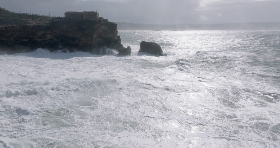 Aerial drone shot of big waves breaking on rocks and cliffs on a day with giant waves in Nazare, Portugal, Europe. Farol da Nazaré lighthouse visible. Shot in ProRes 422 HQ