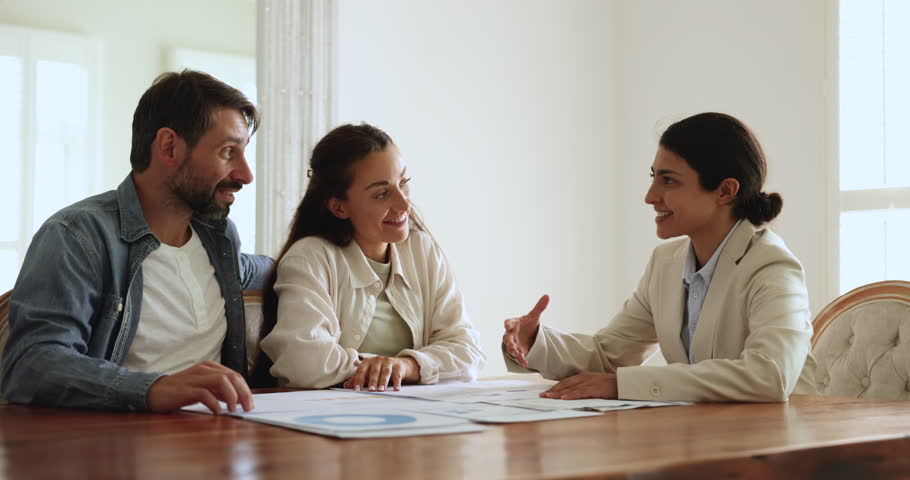 Happy Latin couple meeting with Indian financial advisor, consultant, discussing financial statistic data at table, talking, discussing insurance, investment, planning loan, mortgage, counting profit