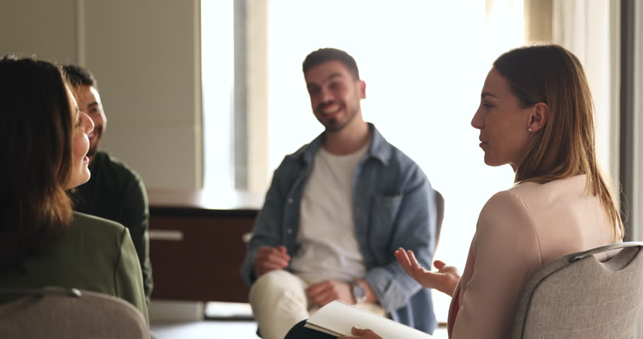 Positive young Caucasian business woman speaking to team of colleagues at teambuilding training meeting, laughing with coworkers. Diverse group talking on therapy session