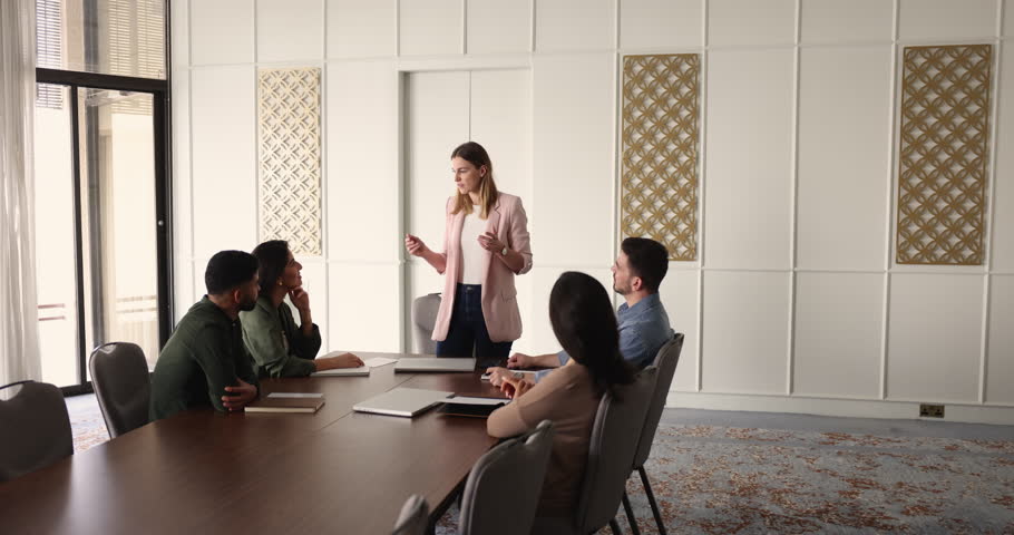 Female manager holding meeting with diverse team, standing at leaders place at large table, talking to colleagues, answering questions, presenting solutions for project. Wide shot with copy space