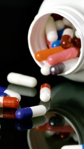 Pill bottle falls over and slips pills onto a black surface in slow motion. Vertical Video.