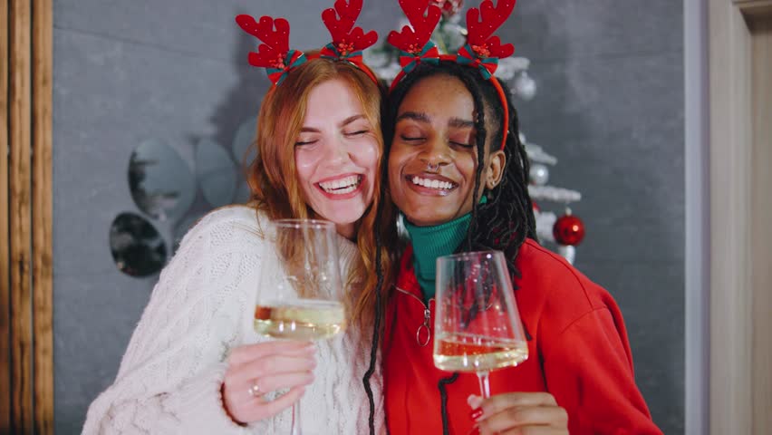 Two joyful women in festive reindeer antlers celebrate with sparkling drinks in front of a beautifully decorated Christmas tree filled with cheerful lights and ornaments