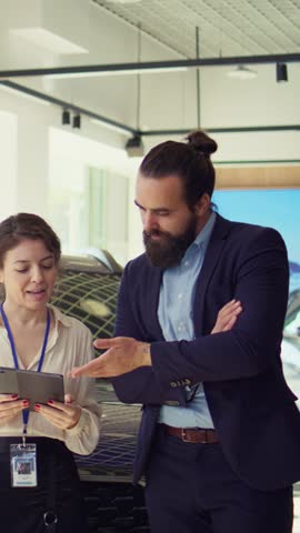 Vertical video Saleswoman using tablet to present car addons options to man, trying sway him to make purchase. Showroom agent offers extended warranty to client, showing details on device, camera B