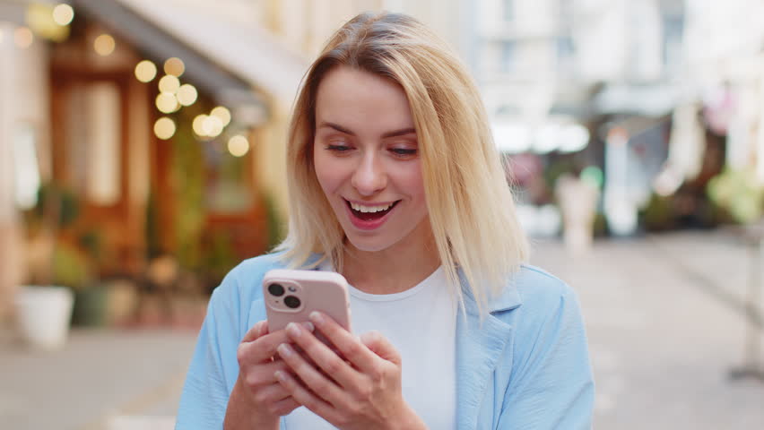 Oh my God Wow. Excited happy joyful winner young woman using smartphone looking at screen browsing celebrating win good message news. Caucasian lady traveler with mobile phone in urban street outdoors