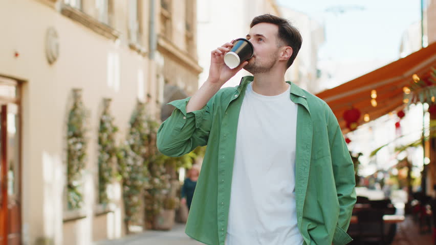 Handsome Caucasian man enjoying morning coffee hot drink and smiling. Relaxing, taking a break. Happy young guy walking on urban city center street, drinking coffee to go. Town lifestyles outside.