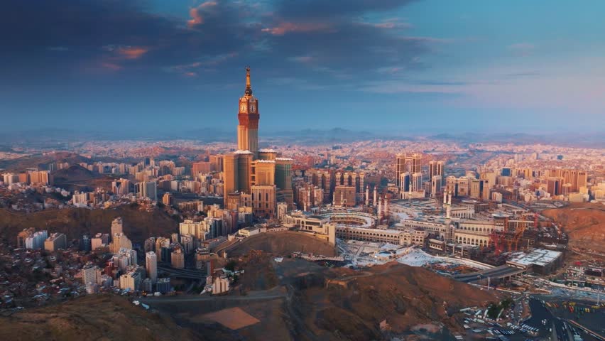 Aerial view of the Abraj Al-Bait Clock Tower and Grand Mosque in Mecca illuminated by the golden light of sunrise, surrounded by the sprawling holy city and mountains