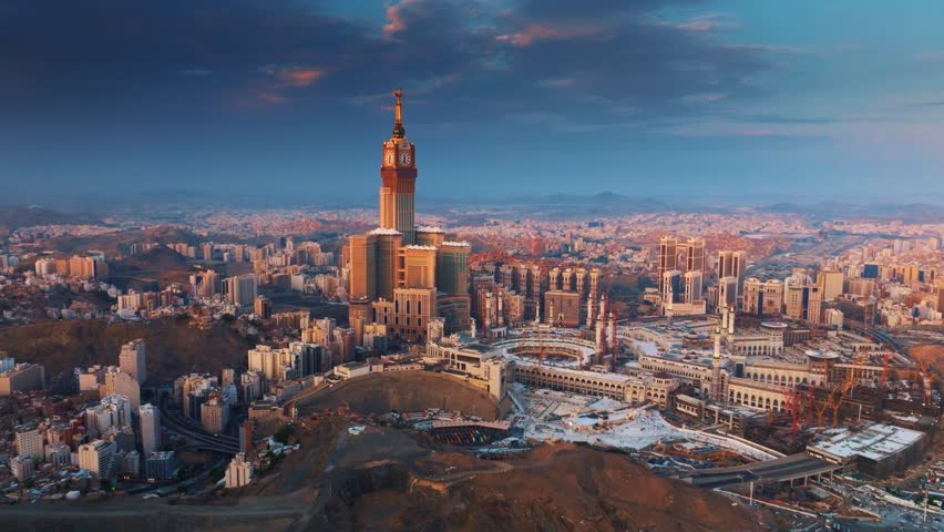 Aerial view of the Abraj Al-Bait Clock Tower and Grand Mosque in Mecca illuminated by the golden light of sunrise, surrounded by the sprawling holy city and mountains