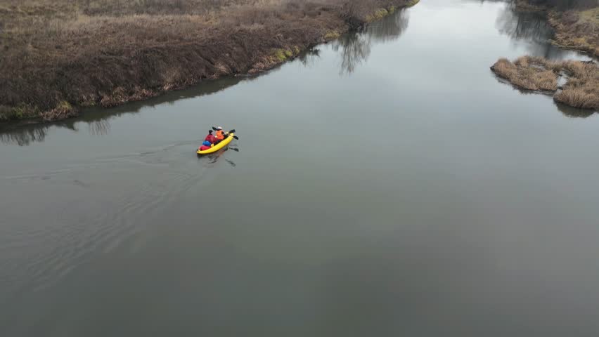 A bright yellow kayak with two rowers slowly sails along the quiet autumn cold river. They row slowly and confidently with oars, guiding the boat along the riverbed. Aerial photography from a drone.