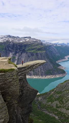 A daring individual stands at the edge of a stunning cliff at Trolltunga, surrounded by majestic mountains and a turquoise fjord below. The beauty of Norway