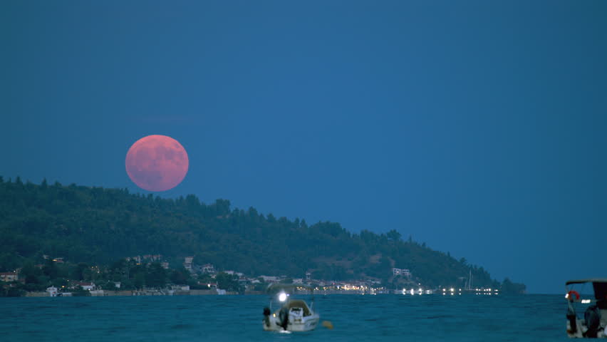 Time lapse of super moon sunset above a tranquil coast, where a small boat drifts in the calm waters, surrounded by a dark blue sky with clouds and distant lights.