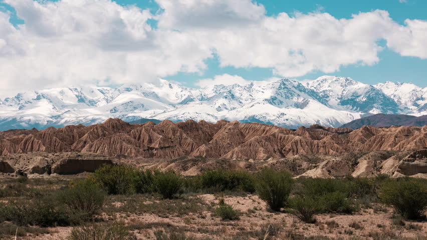 Tien Shan mountain. Kyrgyzstan. Dramatic clouds move over snow-capped mountains, casting shadows on the vibrant red rock formations in the valley, which glow under the warm sunlight of a clear sky