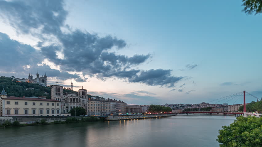 Timelapse of Saint John the Baptist Cathedral and Basilica of Notre-Dame de Fourviere in Lyon, transitioning from day to night. Illuminated waterfront and buildings reflected in the river. France