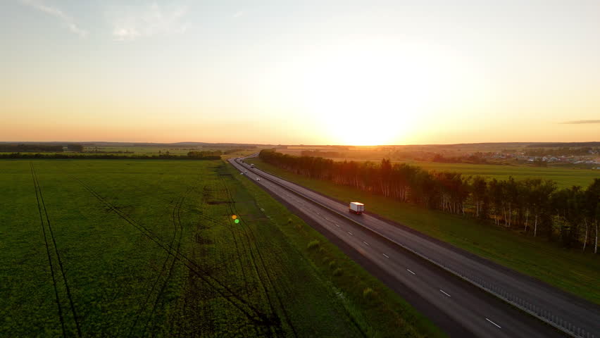 Drone view of the highway in the rays of the setting sun and a moving truck with a semi-trailer. Heavy truck driving on an asphalt road along green fields. Aerial shot