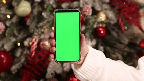Green screen phone in a female hand. Gadget on the background of Christmas tree. Close-up of a female hand and a smartphone with a green screen, New Year. Modern mobile phone. Mockup. Technology - Powered by Shutterstock - Get 15% off with code: PIKWIZARD15