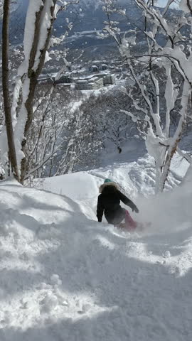 VERTICAL: Female rider in pink pants snowboarding through untouched snow in a dense forest. White powder sprays from board as she navigates between lushly growing snowy trees on a sunny winter day.