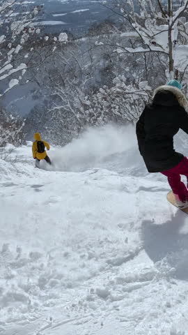 VERTICAL: Two snowboarders carve through fresh powder in a bright, snowy forest. Snow sparkles under sunlight as they search for untouched lines down the mountain. White winter wonderland in Japan.