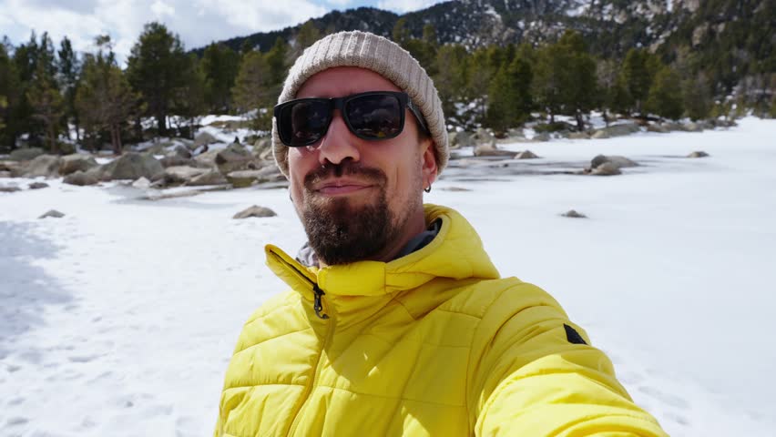 POV of happy Caucasian young man taking selfie video call with mobile in snowy mountains. Millennial handsome male in yellow looking smiling at camera sunny day of winter nature holidays