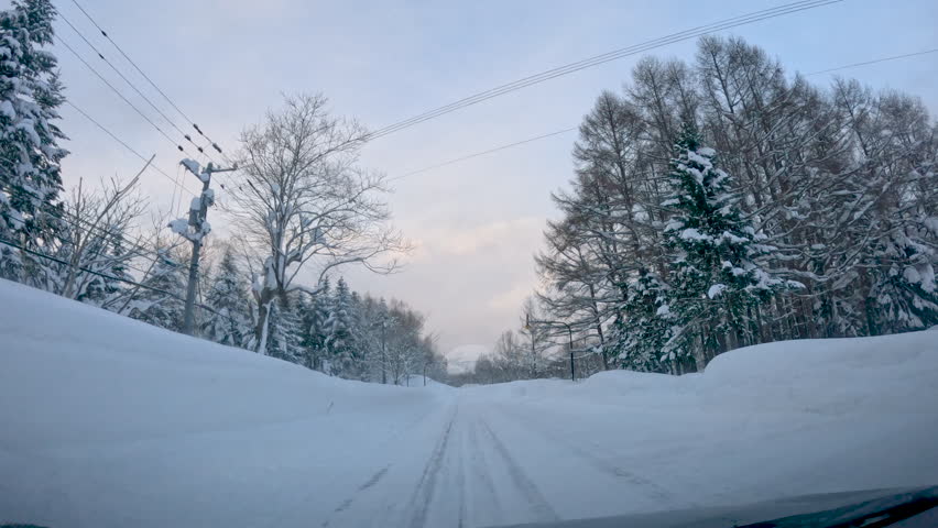 Empty winter road surrounded by towering snowbanks leads towards snowy Mount Yotei. Trees and road are covered with fresh snow, which promises amazing day for powder riding in Japanese mountains.