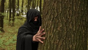 Hooded figure, Hidden figure, Shadowy figure. Mysterious hooded figure with painted hand grips tree trunk in shadowy forest, evoking secrecy and suspense. - Powered by Shutterstock - Get 15% off with code: PIKWIZARD15