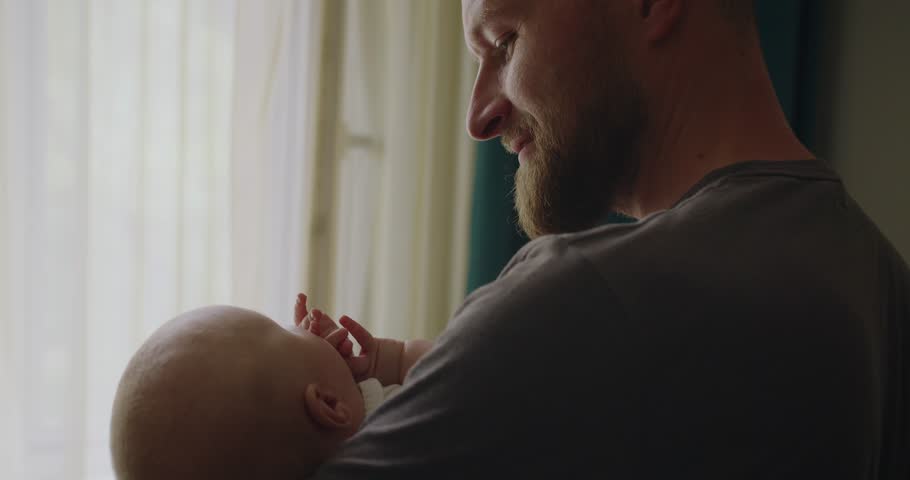 A young father tries to calm and put to sleep a baby holding him in her arms in the children bright room. Affectionate father calming down small babe sing quiet song putting child to sleep.