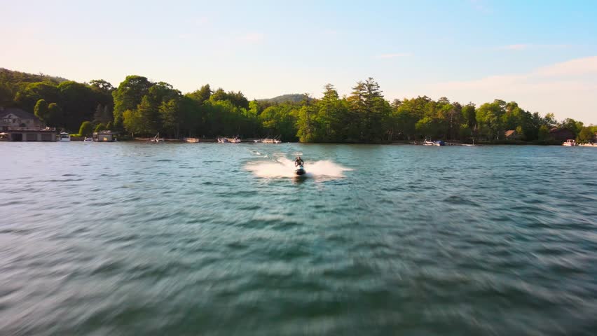 Man riding jetski cinematic aerial chase footage, beautiful Lake George NY backdrop, 4k drone footage, minimal color grade. 
