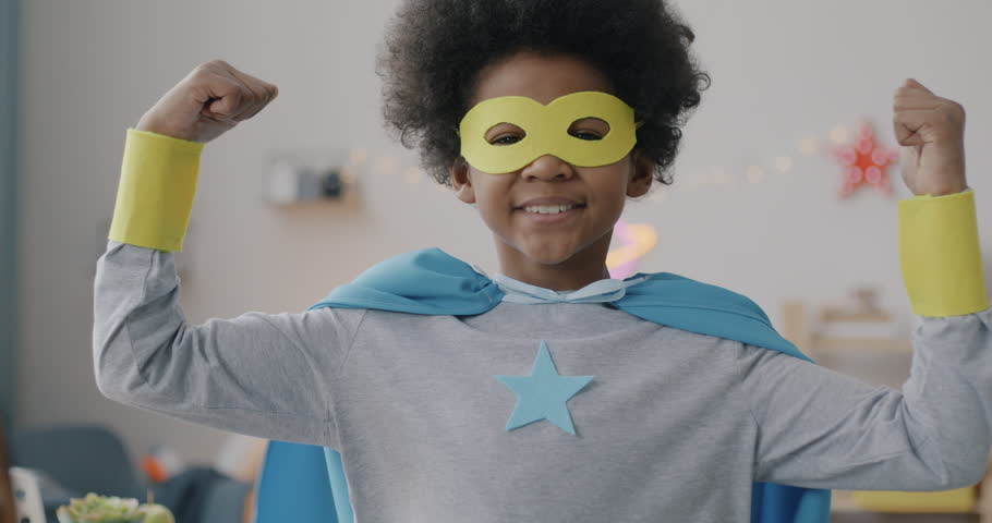 Cute African American child in super hero costume showing biceps and smiling standing indoors at home. Childhood and funny game concept.