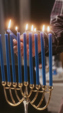 Vertical close up shot of hands of man lighting blue menorah candles while getting ready for Hanukkah holiday at home