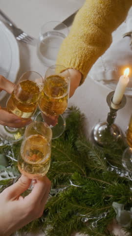 Vertical high angle shot of hands of friends clinking glasses of champagne in toast over festive table decorated with pine branches during Christmas dinner party