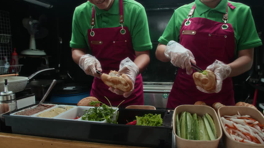 Tilt up shot of cheerful Asian fastfood workers preparing fresh sandwiches with various ingredients in food truck