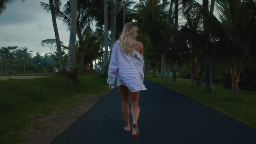 Portrait: Beautiful woman in red swim suit with blonde hair, walking among tropical palm tree. Sexy girl in Asia is flirting in a white shirt. Blonde female enjoys a vacation in bali indonesia