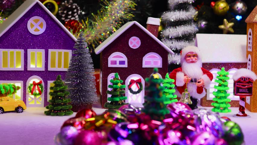 A small Christmas tree surrounded by balls and colorful decorations rotates against the backdrop of a Christmas town with colorful lights in the windows of houses and Santa Claus