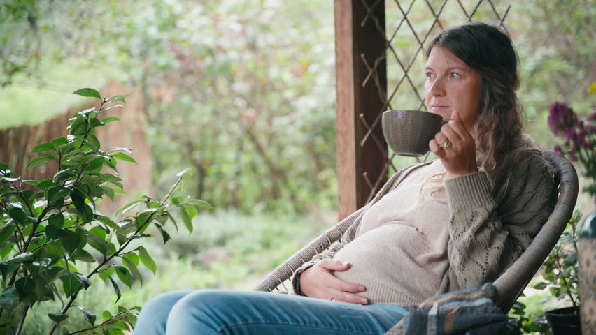 A pregnant middle-aged woman is relaxing and drinking tea while sitting on the terrace of a cottage on a warm autumn day