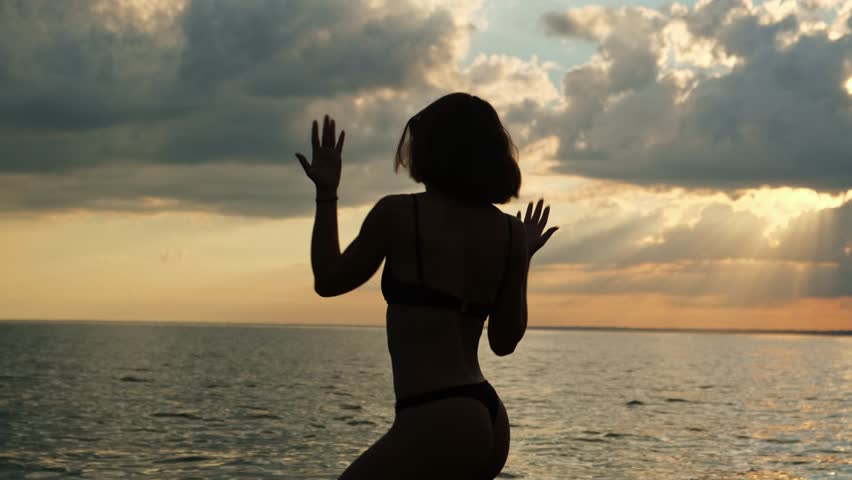 Grace silhouette of young sexy woman in bikini dancing by the sea at sunset. Happy female tourist on the beach enjoying life.
