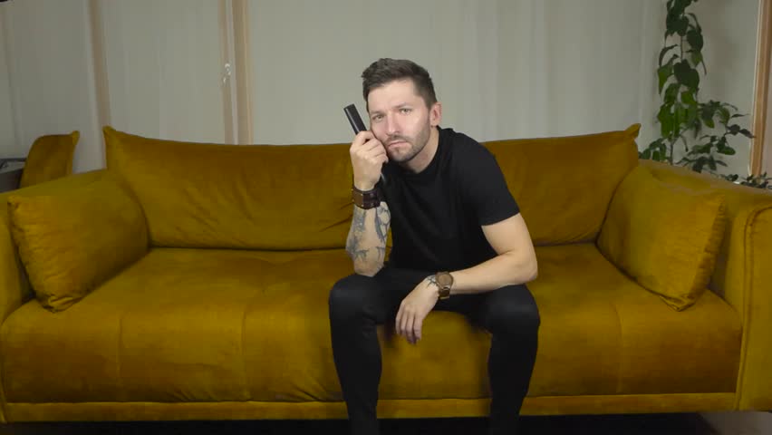 Man watching straight into the camera and pointing tv remote forward to switch channels on the tv that appears to be in front of him. Movement is in slow motion and the man has full sleeve tattoos.