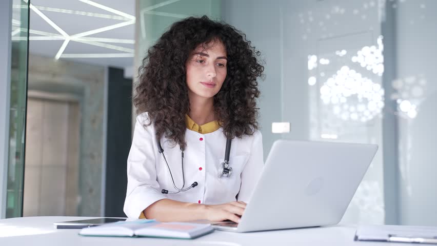 Focused young female doctor is using, typing on a laptop while sitting at a desk at a workplace in hospital clinic. A brunette woman medical worker physician works on a computer in the modern office