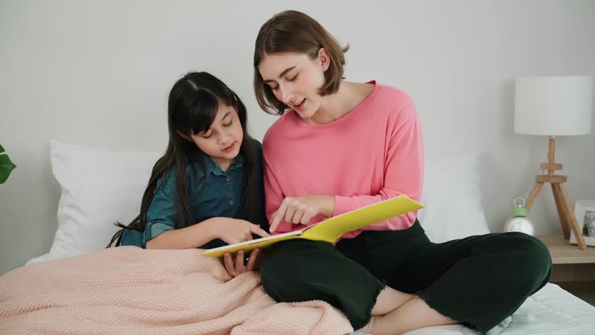 Caucasian mother reading story book to cute daughter listening story. Asian girl pointing while talking about story before bed time. Happy family sitting on bed while spend time together. Pedagogy.