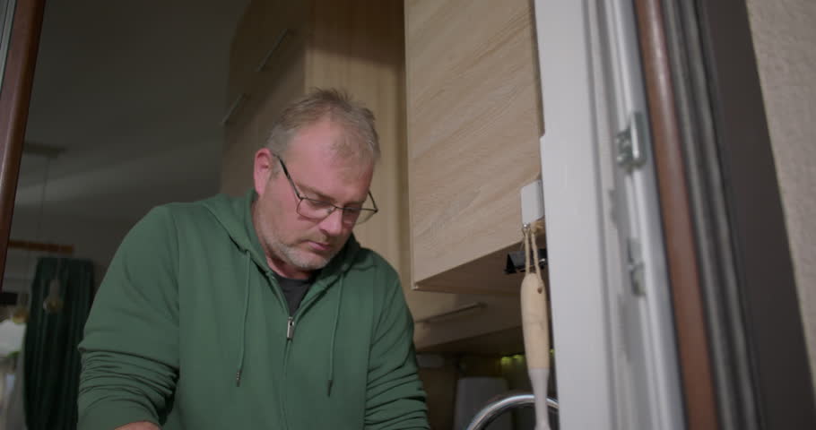 Hands scrubbing a cutting board with a sponge under running tap water at a kitchen sink, depicting daily household cleaning chores in a modern home environment
