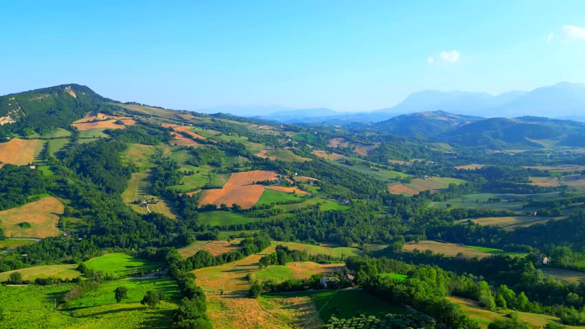 Precious aerial truck left shot from Monte San Martino with checkered fields of green and brown interspersed with vivid green thickets, scattered buildings, amazing Sibillini Mountains, puffy clouds