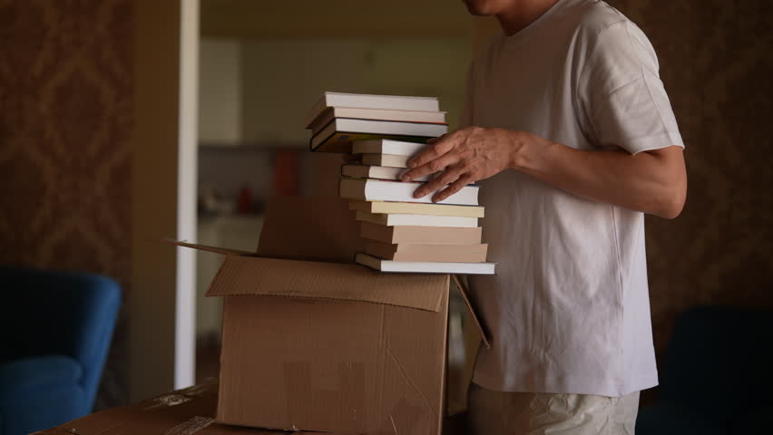 Side view of unrecognizable man packing books into cardboard box, preparing for move, organizing and looking forward to new beginning, close-up, slow motion. Concept of new life in new home relocation