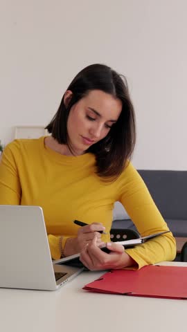 Vertical HD video of young adult student woman taking notes while using laptop computer at home. Millennial pretty female learning online listening virtual video call. Business and education concept